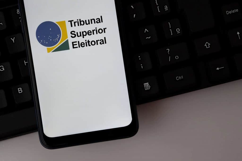 Logotipo do Tribunal Superior Eleitoral (TSE) exibido na tela de um smartphone sobre um teclado de computador.